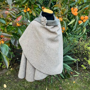 Könnte beinhalten: Ein beigefarbener Wollmantel mit grauem Fleecefutter. Der Mantel hat eine abgerundete Form und ist über einen Mannequin drapiert. Der Mantel hat eine dekorative, blattartige Brosche am Kragen.