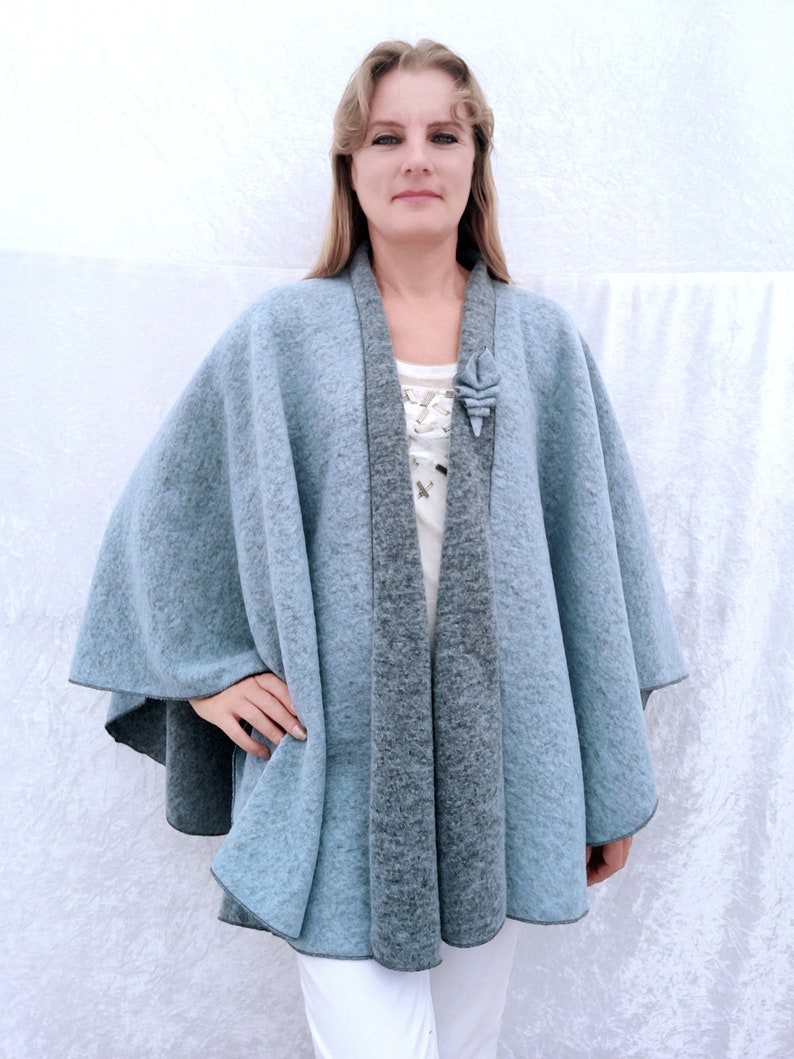 May include: A light blue and grey wool cape with a decorative flower brooch. The cape has a shawl collar and a flowing design.