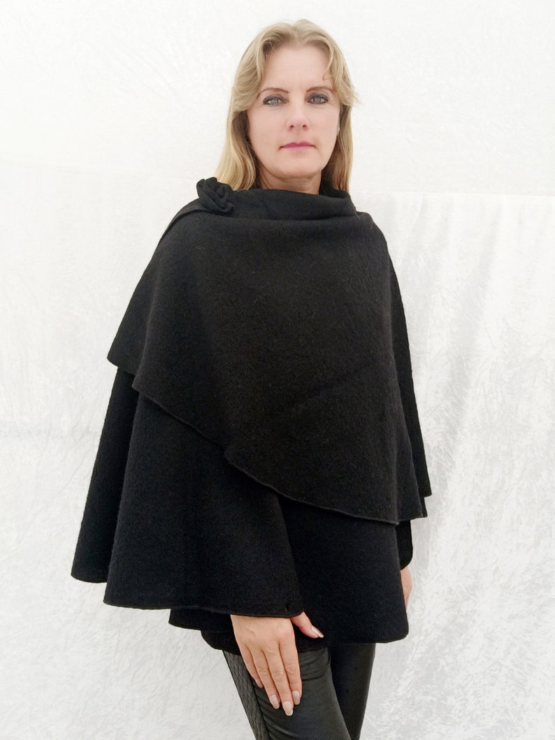 May include: A black wool cape with a cowl neck and a decorative button detail. The cape is draped over the shoulders and falls to mid-thigh.