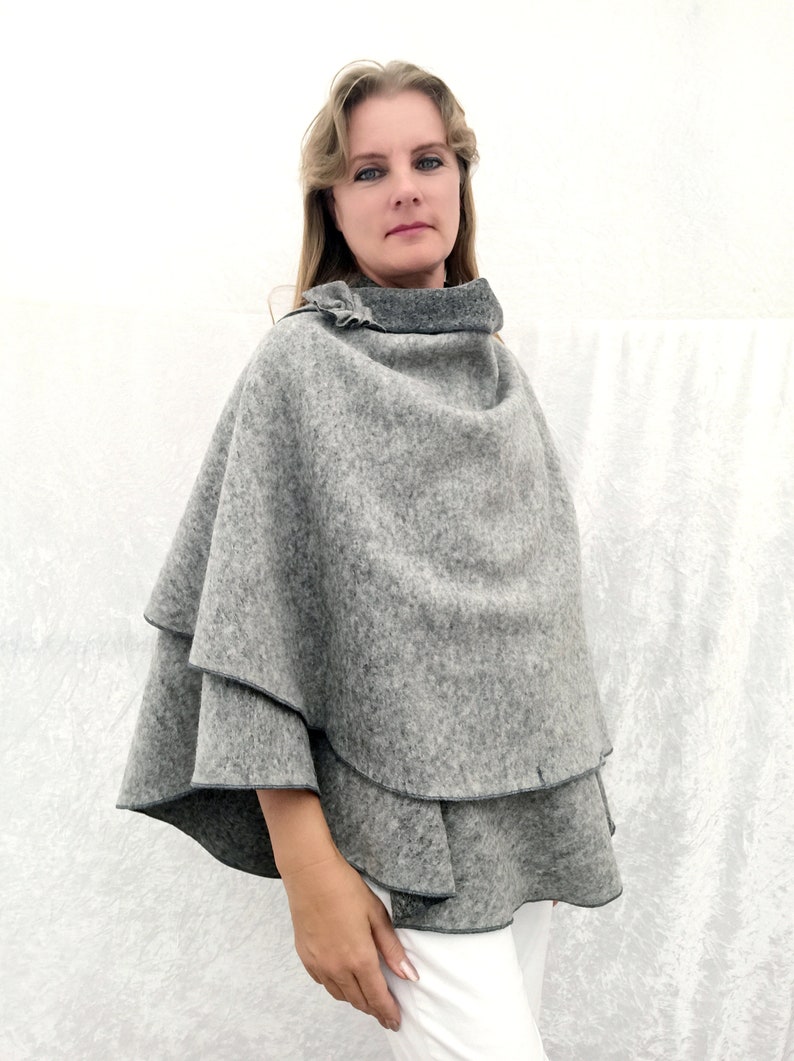 May include: A grey wool poncho with a ruffled neckline. The poncho is layered and has a slightly flared shape.