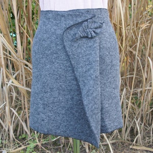 Könnte beinhalten: Ein grauer Wickelrock mit einem dekorativen Detail auf der Vorderseite. Der Rock ist aus einem weichen, strukturierten Stoff.