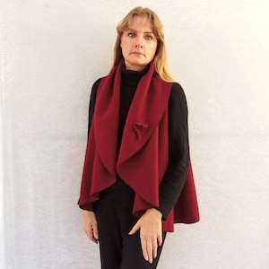 Könnte beinhalten: Eine burgunderfarbene, ärmellose Weste mit einem breiten, drapierten Kragen und einer dekorativen Brosche. Die Weste ist aus einem weichen, strukturierten Stoff gefertigt.