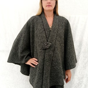May include: A grey wool cape with a knotted tie closure. The cape has a wide, flowing silhouette and a simple, elegant design.