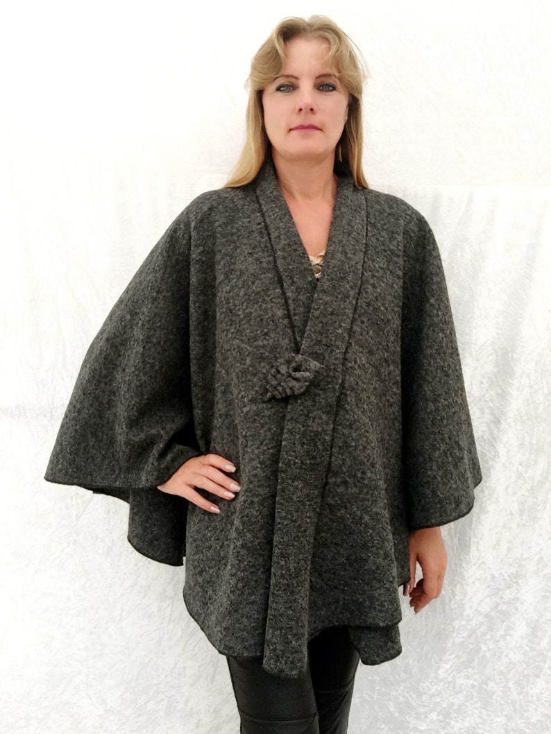 May include: A grey wool cape with a knotted tie closure. The cape has a wide, flowing silhouette and a simple, elegant design.