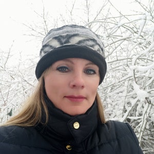 May include: A woman wearing a black and white patterned hat with a black brim and a black jacket. The background features snow-covered branches, creating a winter scene. The hat is the focal point of the image.