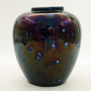 May include: A round, iridescent ceramic vase with a dark blue rim. The vase has a glossy finish with a blend of dark green, purple, and bronze hues. Small, light blue spots are scattered across the surface.