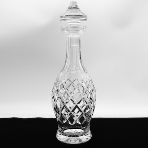 May include: A clear, cut-glass decanter with a diamond pattern and a conical stopper. The decanter has a long neck and a bulbous body, and sits on a round base. The stopper is also cut glass.