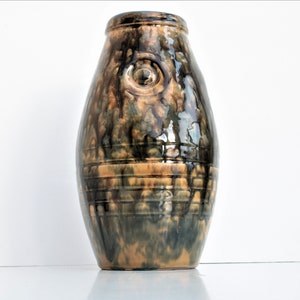 May include: A brown and green ceramic vase with a textured surface and a raised circle design. The vase has a rounded, oval shape.