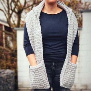 May include: A gray knitted scarf with two pockets. The scarf is worn by a person wearing a dark blue shirt and blue jeans.