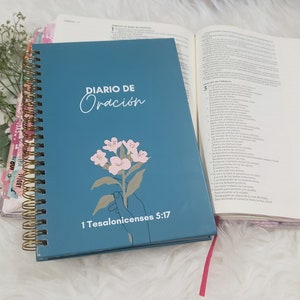 Puede incluir: Un diario de oración azul turquesa con una encuadernación en espiral dorada. La portada presenta una ilustración de una flor rosa y el texto "DIARIO DE ORACION" y "1 Tesalonicenses 5:17".
