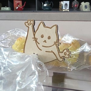 May include: A light-colored wooden cat-shaped clip with a laser-engraved design of a cat with one paw raised. The clip is attached to a clear plastic bag containing baked goods, featuring a simple outline of a cat face, whiskers, and paws.