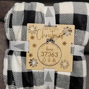 May include: A black and white buffalo plaid blanket with a wooden sign that says "Wishing you a warm and cozy Christmas. There's no place like home for the holidays." The sign also has a snowflake and a Christmas tree design.