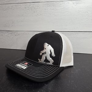 May include: Black and white trucker hat with a Bigfoot silhouette embroidered on the front. The hat features a black front panel, a white mesh back, and a curved brim with white stitching. The hat is adjustable.