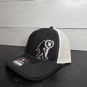 May include: Black and white trucker hat with a Bigfoot graphic holding a TN highway sign. The hat features a black front panel, white mesh back, and a curved brim. The Richardson brand label is visible.