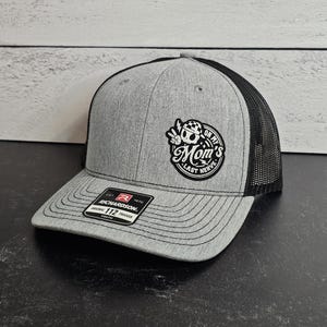 May include: Gray trucker hat with a black mesh back. The hat features a patch with the text "Mom's Last Nerve" and a skull graphic. The brim is curved, and the hat has a Richardson tag.