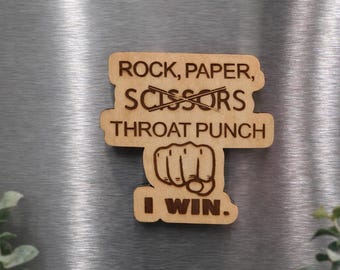 May include: A wooden magnet with the text "ROCK, PAPER, SCISSORS, THROAT PUNCH, I WIN." The word "scissors" is crossed out, and a fist graphic is below "THROAT PUNCH."
