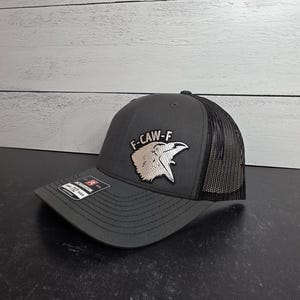 May include: A charcoal gray trucker hat with a black mesh back. The hat features the text "F-CAW-F" above a silver bird graphic. The hat has a curved brim and a Richardson 112 tag.