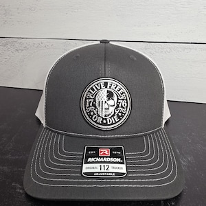 May include: A gray trucker hat with a white mesh back and a curved brim. The front features a circular patch with a skull design and the words "LIVE FREE OR DIE". The hat has a Richardson tag and is adjustable.