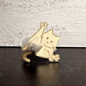 May include: A wooden cat-shaped ornament with a paw raised in the air. The cat is light brown with gold-colored detailing. The ornament is sitting on a dark surface with a white wooden background.