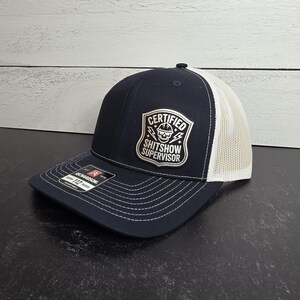 May include: Navy blue and white trucker hat with a curved brim. The front features a patch with the words "CERTIFIED SHITSHOW SUPERVISOR" and a skull graphic. The hat has a mesh back and adjustable snap closure.