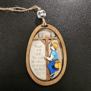 May include: Wooden ornament in an oval shape, featuring a lineman on a utility pole. The ornament has a light brown wooden frame, with the text "When God said, 'let there be light...' he created linemen."