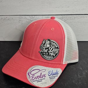 May include: A coral-colored baseball cap with a white mesh back. The cap features a circular patch with the text "Have The Day You Deserve" and a skeleton graphic. The hat also has a tag that says "Evolve Elude".