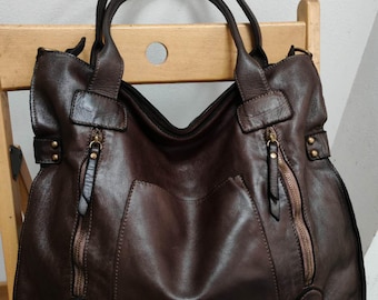 May include: A dark brown leather handbag featuring two top handles, a central open pocket, and two vertical zippered pockets on the front. The bag includes brass-toned hardware, decorative side straps with metal studs, and D-rings at the base.