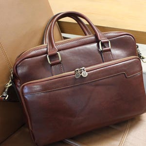 May include: A brown leather briefcase with a top handle and a zippered front pocket. The briefcase has a gold-toned zipper and hardware.