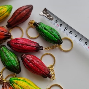 Könnte beinhalten: Eine Sammlung von handbemalten, bunten Kakaoschoten-Schlüsselanhängern. Jeder Schlüsselanhänger hat eine andere Farbkombination, darunter Rot, Grün und Gelb. Die Schlüsselanhänger sind etwa 5 cm lang und an goldfarbenen Schlüsselringen befestigt.
