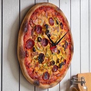 May include: A pizza-shaped wall clock with a realistic design. The clock features pepperoni, olives, and other toppings on a red sauce base. Black clock hands indicate the time. The clock is mounted on a white wooden wall.