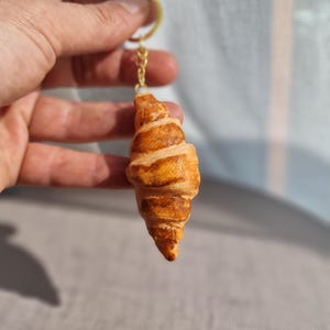 Op de afbeelding: Een gouden sleutelhanger met een realistische croissant-hanger. De croissant is lichtbruin van kleur met een bladerige textuur.