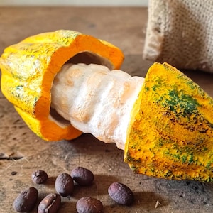 Puede incluir: Una mazorca de cacao partida, amarilla y verde, que revela la pulpa blanca y los granos de cacao. Varios granos de cacao de color marrón oscuro están esparcidos sobre una superficie de madera. Una bolsa de arpillera está en el fondo.