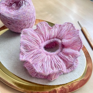 May include: A pink and white yarn hair scrunchie with a ruffled edge. The scrunchie is on a gold plate with a white background. A ball of pink and white yarn is in the background.