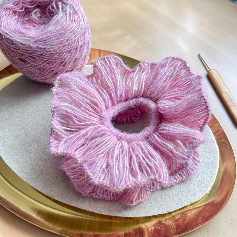 May include: A pink and white yarn hair scrunchie with a ruffled edge. The scrunchie is on a gold plate with a white background. A ball of pink and white yarn is in the background.