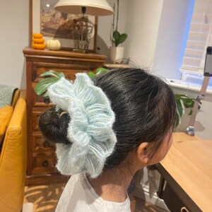 May include: A light blue fuzzy scrunchie with a gathered, ruffled design. The scrunchie is worn in a person's hair.