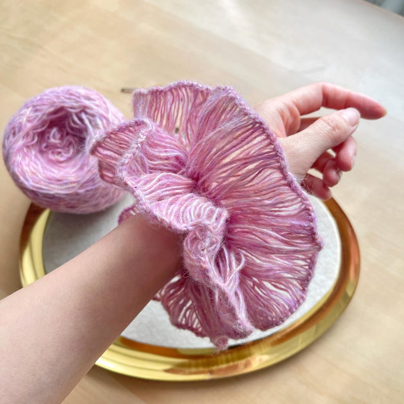 May include: A pink crocheted ruffle bracelet with a delicate, lacy design. The bracelet is made with a soft, fluffy yarn and has a slightly textured appearance.