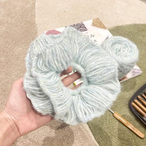May include: A light blue and white yarn hair scrunchie, crocheted in a ruffled style. The scrunchie is being held in a hand, with a ball of yarn and crochet hooks visible in the background.
