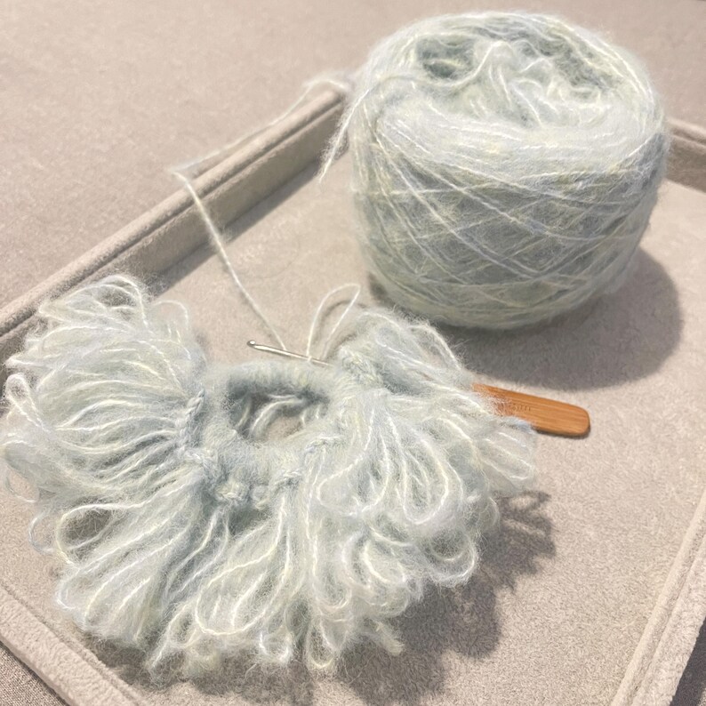 May include: A ball of light blue and white yarn with a crochet hook and a partially crocheted piece of fabric.
