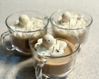 Könnte beinhalten: Drei transparente Glasbecher gefüllt mit einer hellbraunen Flüssigkeit, bedeckt mit weißer Schlagsahne und einem Schneemann-Marshmallow. Die Schneemänner haben kleine orangefarbene Nasen und schwarze Knöpfe. Die Becher haben transparente Griffe.