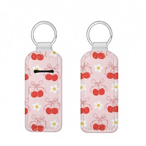 May include: Two keyrings with a pink background and a repeating pattern of red cherries tied with bows and white daisies. Each keyring has a silver ring at the top.