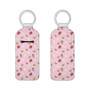 May include: A pink keychain case with a strawberry pattern. The case has a repeating design of red strawberries, white flowers, and green leaves on a striped background. A silver key ring is attached to the top.