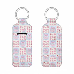 Peut inclure: Deux porte-clés avec un motif répété de cerises roses et de carrés vichy bleus sur fond blanc. Les porte-clés ont un anneau argenté en haut et une fente noire sur un côté.