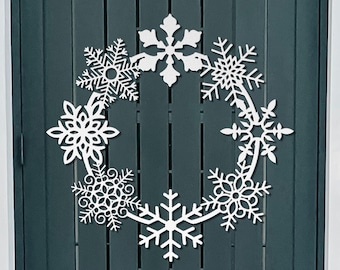 May include: A white snowflake wreath made of cut-out snowflakes hangs on a dark gray wooden door. The wreath is made of six snowflakes, each with a different design. The door is made of vertical wooden planks.