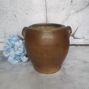 May include: A brown ceramic jar with two handles. The jar has a rounded body and a slightly flared top. The jar is sitting on a gray surface.