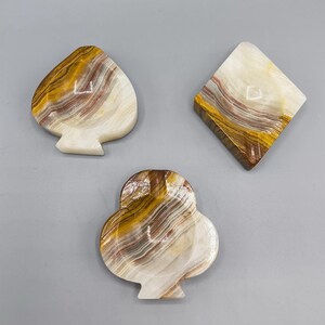 Op de afbeelding: Drie decoratieve marmeren asbakken in de vorm van speelkaartsymbolen: schoppen, klaveren en ruiten. De asbakken zijn gemaakt van wit marmer met bruine en rode strepen.