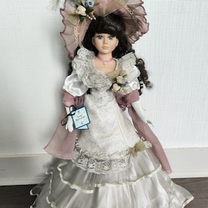 Puede incluir: Muñeca de porcelana con un largo vestido blanco en capas con detalles de encaje y cintas. La muñeca tiene cabello oscuro, ojos azules y un collar rosa. Lleva un sombrero grande y adornado con plumas y flores.