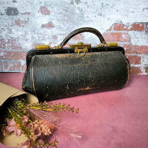 May include: A vintage black leather doctor's bag with brass hardware and a worn, distressed look. The bag is sitting on a pink surface with a bouquet of pink flowers in the foreground.