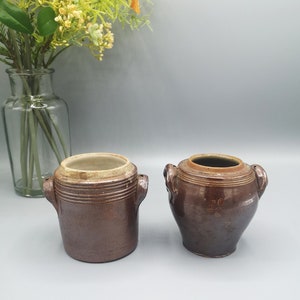 Op de afbeelding: Twee bruine keramische kruiken met handvatten. De kruik links is cilindrisch en heeft een iets lichtere bruine bovenkant. De kruik rechts is ronder en heeft een donkerdere bruine glazuur. Beide kruiken hebben een rustieke, vintage uitstraling.