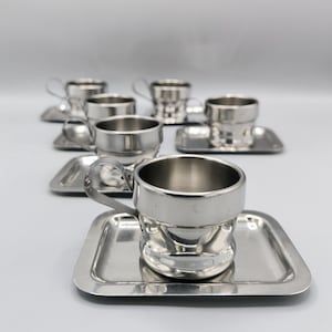 Può includere: Un set di sei tazzine da espresso e piattini d'argento. Le tazzine sono piccole e rotonde, con un manico su un lato. I piattini sono quadrati e hanno un bordo leggermente rialzato.