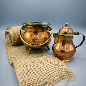 Puede incluir: Una olla de cobre con acabado martillado y asas de latón se encuentra sobre un corredor de arpillera con una tetera de cobre a su lado.